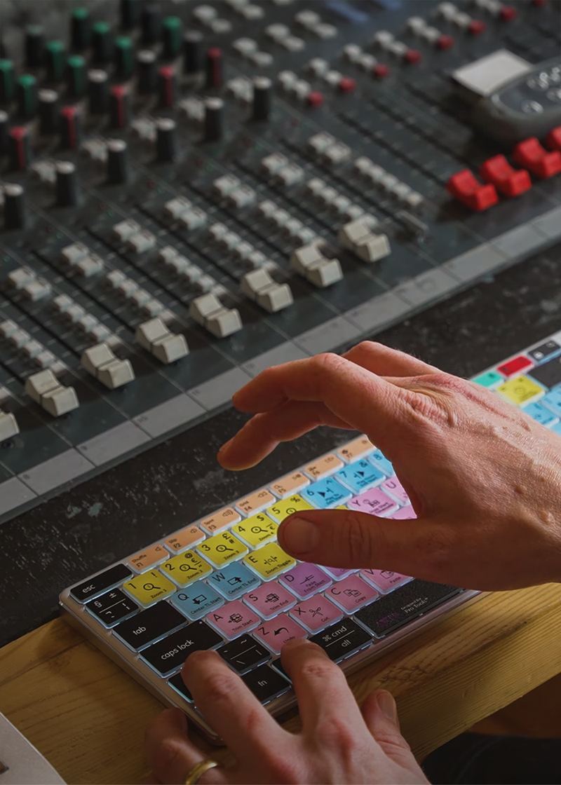A close-up of a person's hands using a colorful keyboard overlay on a computer keyboard in front of a large audio mixing board from Editors Keys 